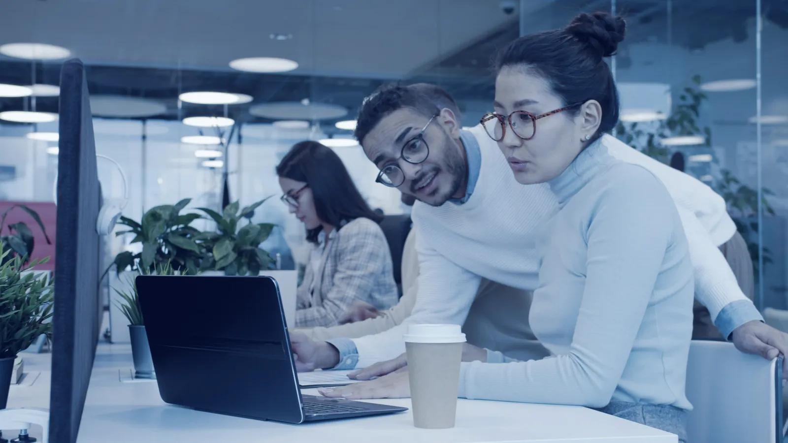 Two colleagues leaning over a laptop together in an open office, talking through what they see